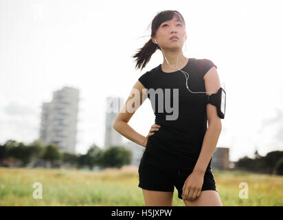 Colpo all'aperto del femminile in piedi nel parco urbano. Donna cinese in sportswear con gli auricolari per ascoltare musica e guardare Foto Stock