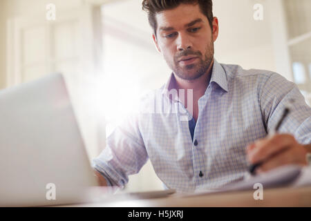 Colpo di giovane imprenditore seduti a casa ufficio con computer portatile e prendere appunti. Giovane uomo che lavora da casa. Foto Stock