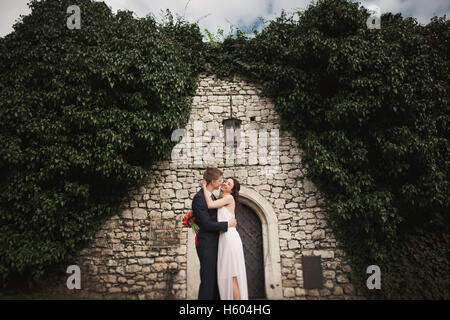 Splendida sposa in posa vicino al bellissimo muro di cespugli di piante di alberi nel loro giorno di nozze Foto Stock