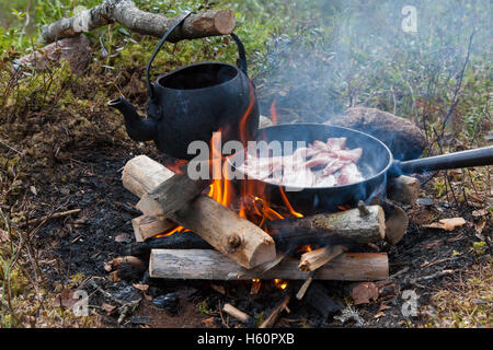 Stagno annerito bollitore di acqua bollente e in padella la pancetta su fiamme dal fuoco durante il trekking nella foresta Foto Stock