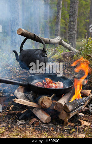 Stagno annerito bollitore di acqua bollente e in padella la pancetta su fiamme dal fuoco durante il trekking nella foresta Foto Stock