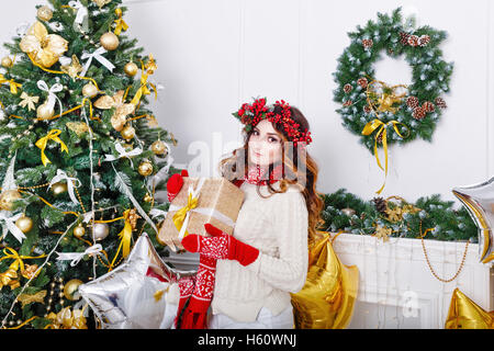 Bella ragazza in una sciarpa e guanti tenendo un dono in un albero di Natale. L'atmosfera di festa. Foto Stock