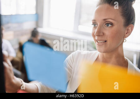 Giovane donna che guarda su un post-it e di parete di brainstorming. Imprenditrice in piedi presso l'ufficio dietro la parete in vetro con STIC Foto Stock