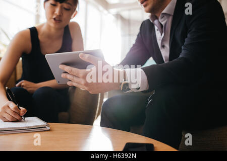 Immagine ritagliata di imprenditore azienda digitale compressa e la donna con il blocco note. Gli imprenditori seduti nella lobby e lavorare insieme. Foto Stock