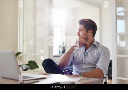 Colpo di giovane uomo seduto a tavola che guarda lontano e di pensare. Imprenditore pensieroso seduta home office. Foto Stock