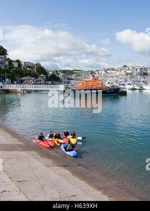 I giovani imparano a canoa in Brixham Harbour, devon con il rnli scialuppa di salvataggio in background. Foto Stock