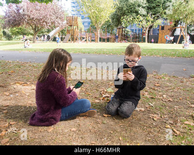 Due bambini di giocare i giochi su telefoni cellulari in un parco pubblico con un'area di gioco dietro di loro. Foto Stock