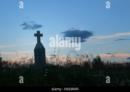 Una vecchia croce headstone silhouette in un campo erboso con Fiori al tramonto Foto Stock