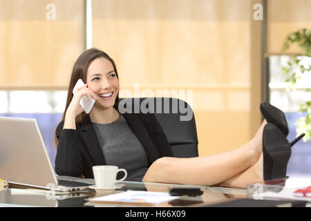 Rilassata imprenditrice chiamando al telefono seduto in una sedia con le gambe su un desktop in ufficio Foto Stock