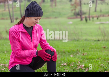 Giovane atleta femminile male da un infortunio al ginocchio in una fredda giornata invernale sulla via di un parco urbano. Foto Stock