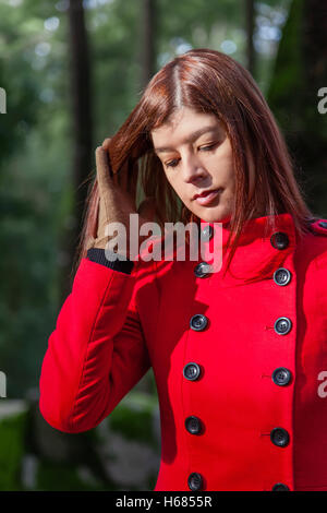 Giovane donna triste sensazione di camminare su una foresta che indossa un cappotto rosso in inverno sotto un raggio di luce solare Foto Stock