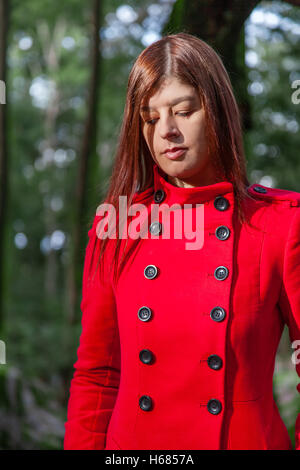 Giovane donna triste sensazione di camminare su una foresta che indossa un cappotto rosso in inverno sotto un raggio di luce solare Foto Stock