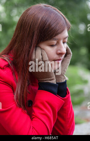 Ritratto di una giovane donna depressa seduto su una foresta che indossa un soprabito rosso durante il periodo invernale Foto Stock