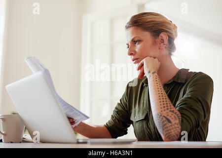 Colpo di giovane donna alla lettura di documenti mentre si lavora sul computer portatile. Donna seduta a tavola in ambienti chiusi e la lettura di alcuni documenti. Foto Stock