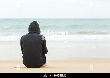 Vista posteriore del ritratto di un ragazzo adolescente pensando solo e guardando il mare seduti sulla sabbia della spiaggia Foto Stock