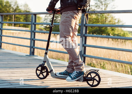 Giovane uomo in abbigliamento casual su kick scooter in parco al tramonto Foto Stock