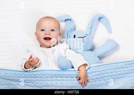 Funny little baby indossando una calda giacca a maglia giocando con il giocattolo bunny rilassante sul cavo bianco coperta di maglia nella soleggiata vivaio. Foto Stock