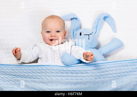 Funny little baby indossando una calda giacca a maglia giocando con il giocattolo bunny rilassante sul cavo bianco coperta di maglia nella soleggiata vivaio. Foto Stock