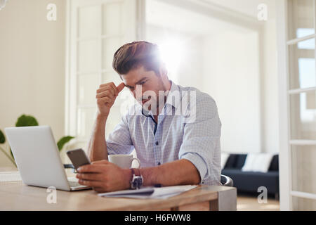 Colpo di giovane serio utilizzando il telefono cellulare mentre è seduto alla sua scrivania. Uomo d'affari a tavola con laptop e guardando sm Foto Stock