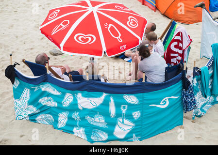 Famiglia come vacanzieri su St Ives beach seduto e parlando dietro un colorato e frangivento parasol su un nuvoloso e ventoso giorno di estate, Cornwall, Regno Unito Foto Stock