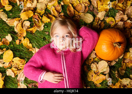 Bambino carina ragazza bionda giacente in foglie di autunno con la zucca. Foto Stock