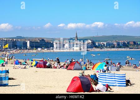 Vista lungo la spiaggia verso il lungomare edifici con vacanzieri godere il sole, Weymouth Dorset, Inghilterra, Regno Unito. Foto Stock