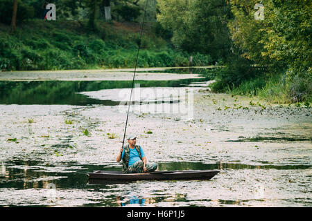 Lone Senior uomo Con Fish-Rod pescatore, seduti in un piccolo canotto in legno in verde fiume sovradimensionate backwaters di stagno in estate. Foto Stock