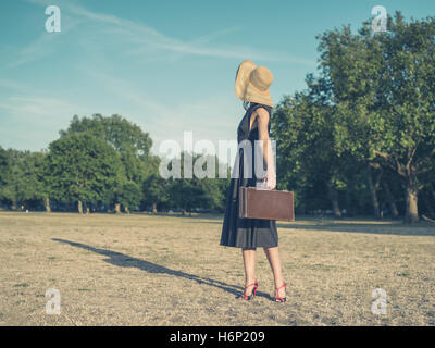 Vintage filtrata colpo di un elegante giovane donna che indossa un vestito e un cappello in piedi nel parco con una valigetta Foto Stock