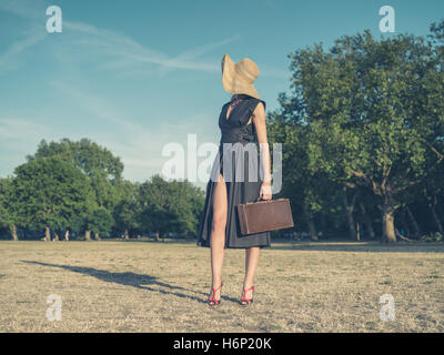 Vintage filtrata colpo di un elegante giovane donna che indossa un vestito e un cappello in piedi nel parco con una valigetta Foto Stock