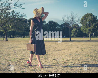 Vintage filtrata colpo di un elegante giovane donna in tacchi alti e un abito a piedi nel parco con una valigetta Foto Stock