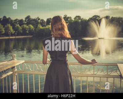 Vintage filtrata colpo di un elegante giovane donna in piedi da un lago in un parco al tramonto Foto Stock