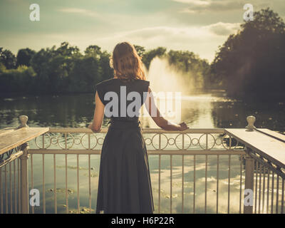 Vintage filtrata colpo di un elegante giovane donna in piedi da un lago in un parco al tramonto Foto Stock