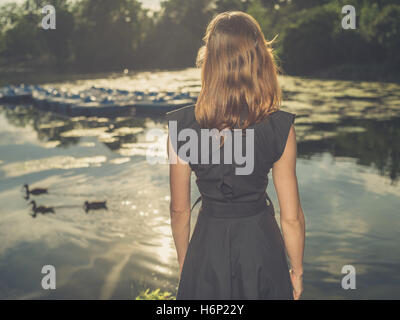 Vintage filtrata colpo di un elegante giovane donna in piedi da un lago in un parco al tramonto Foto Stock