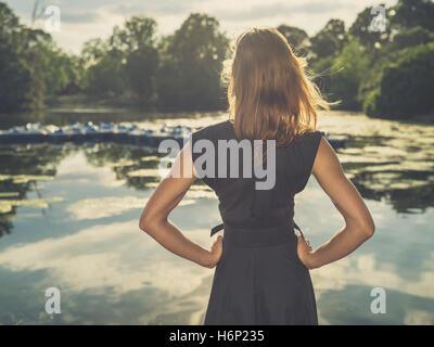 Vintage filtrata colpo di un elegante giovane donna in piedi da un lago in un parco al tramonto Foto Stock