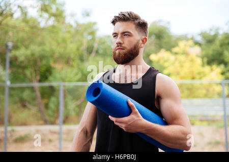 Ritratto di un concentrato barbuto uomo sportive azienda materassino yoga all'aperto Foto Stock