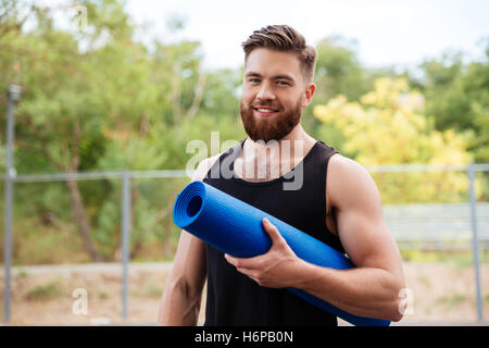 Bello barbuto giovane atleta azienda materassino yoga e guardando la fotocamera all'aperto Foto Stock