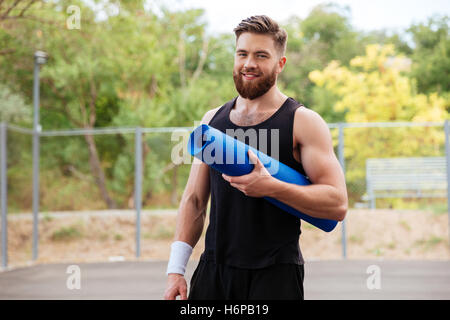 Sorridendo allegro barbuto uomo fitness con materassino yoga in piedi all'aperto Foto Stock