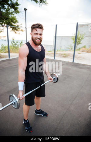 Attraente sportivo sorridente e permanente tenendo barbell all'aperto Foto Stock