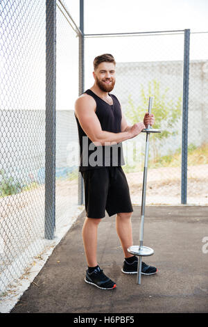 A piena lunghezza Ritratto di un bel giovane sorridente uomo fitness azienda barbell all'aperto Foto Stock