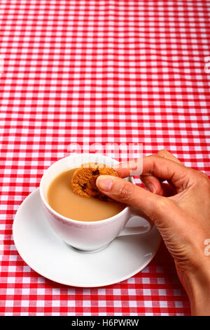 Donna-ladies-ragazze dunking mano un cookie biscotto in una calda tazza di tè con un punto rosso e bianco a scacchi tabella coperta di stoffa Foto Stock