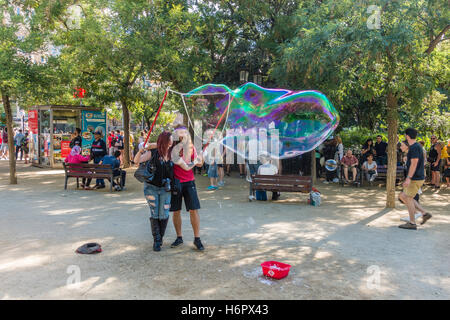 Un giovane di venti-qualcosa di donna spagnola è aiutato da un uomo in enormi bolle di sapone in un parco dalla Sagrada Familia Foto Stock