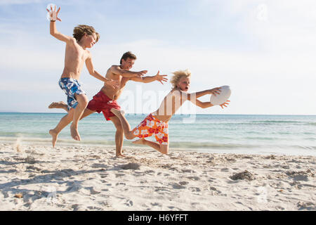 Ragazzo e padre a caccia di fratello con pallone da rugby sulla spiaggia, Maiorca, SPAGNA Foto Stock