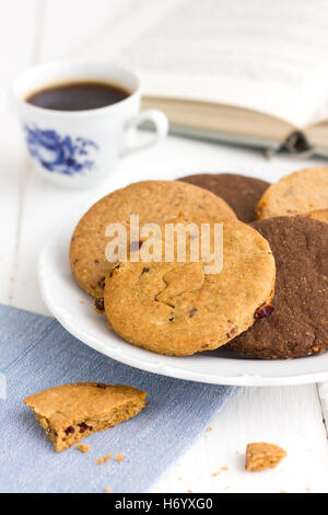 Mescolati i biscotti al cioccolato su una piastra su un tavolo rustico con caffè nero e il libro aperto in background. Foto Stock
