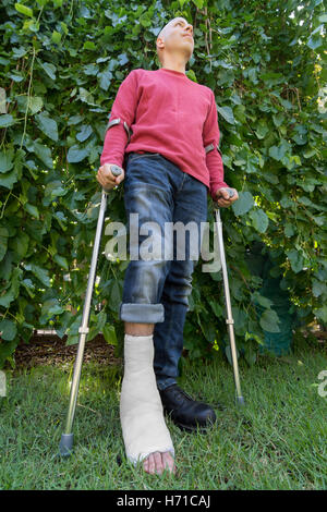 Giovane con una caviglia rotta e una fibra di vetro bianco e il calco in gesso sulla sua gamba, ottenendo un po' di aria fresca in giardino mentre wal Foto Stock