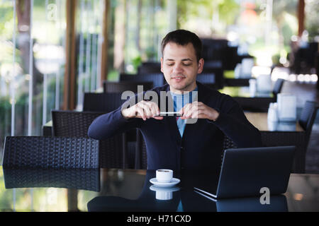 Bel giovane uomo che fa la foto del suo caffè caldo bere utilizzando uno smartphone e sorridenti mentre è seduto nella caffetteria con il computer portatile Foto Stock