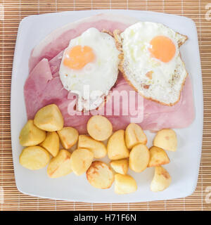 Due uova fritte con fette di prosciutto, patate, su una piastra bianca su una stuoia di vimini visto dal di sopra. Foto Stock