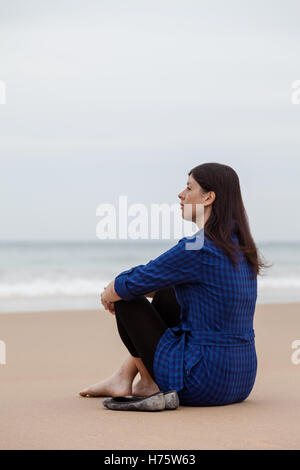 Solitaria e premuto donna seduta sulla sabbia di una spiaggia deserta e guardare il mare su una giornata autunnale. Foto Stock