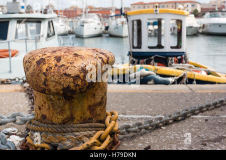 Primo piano della fune e cavo metallico legato al giallo arrugginito Bollard, barche da pesca in background Foto Stock