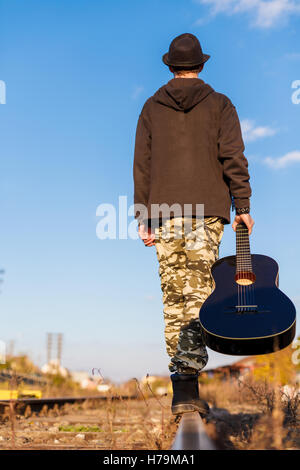 Giovane uomo tenendo la chitarra nelle sue mani è a piedi in un percorso ferroviario in cerca di libertà. concetto musicale, viaggiatore Foto Stock