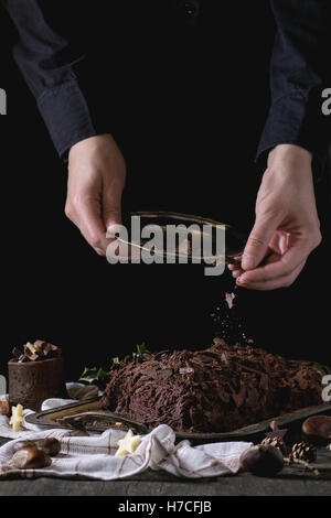 Processo di decorazione di Natale fatti in casa il cioccolato yule log da donna con le mani in mano con scaglie di cioccolato su un vecchio tavolo di legno con hol Foto Stock